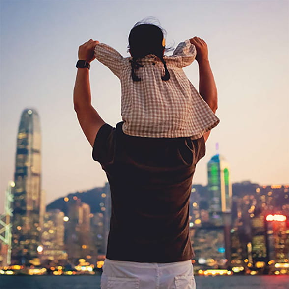 Man facing a city landscape at twilight with a child sitting on his shoulders.