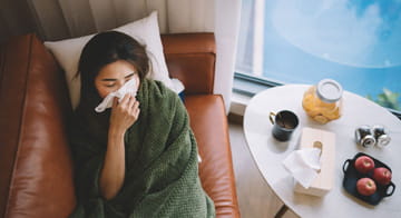 Young woman, wrapped in a blanket on the sofa, feeling unwell and holding a tissue to her nose.