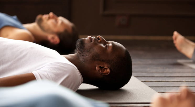 Male lying down on a yoga mat, with his eyes closed, in the middle of a yoga class