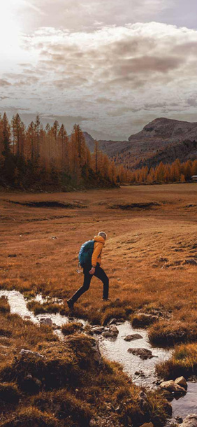 man crossing a river on a hike with mountains in the background