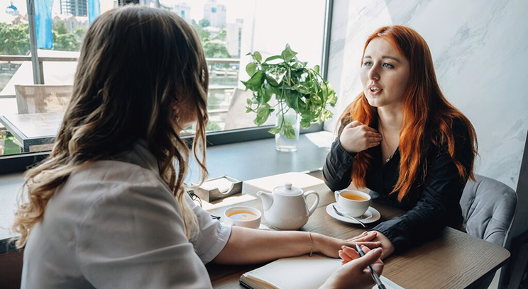 Two women talking in café touching hands. One has pen and notepad