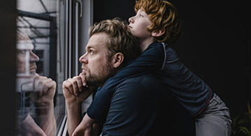 Male adult with young child holding onto his back, both looking contemplatively out of the window.